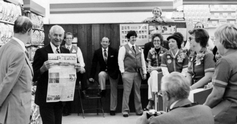 Sam Walton holding up a newspaper of a Walmart store's grand opening, while gathering with group of Walmart employees