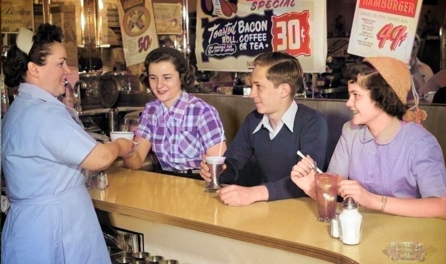 1950s Anonymous Smiling Woman Waitress Serving Two Teenage Girls And One Boy At Diner Lunch Soda Fountain Counter.