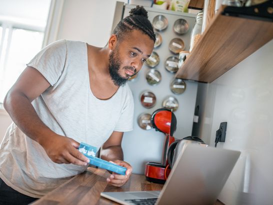 Hispanic man using laptop in the kitchen and holding a pill box in his hands