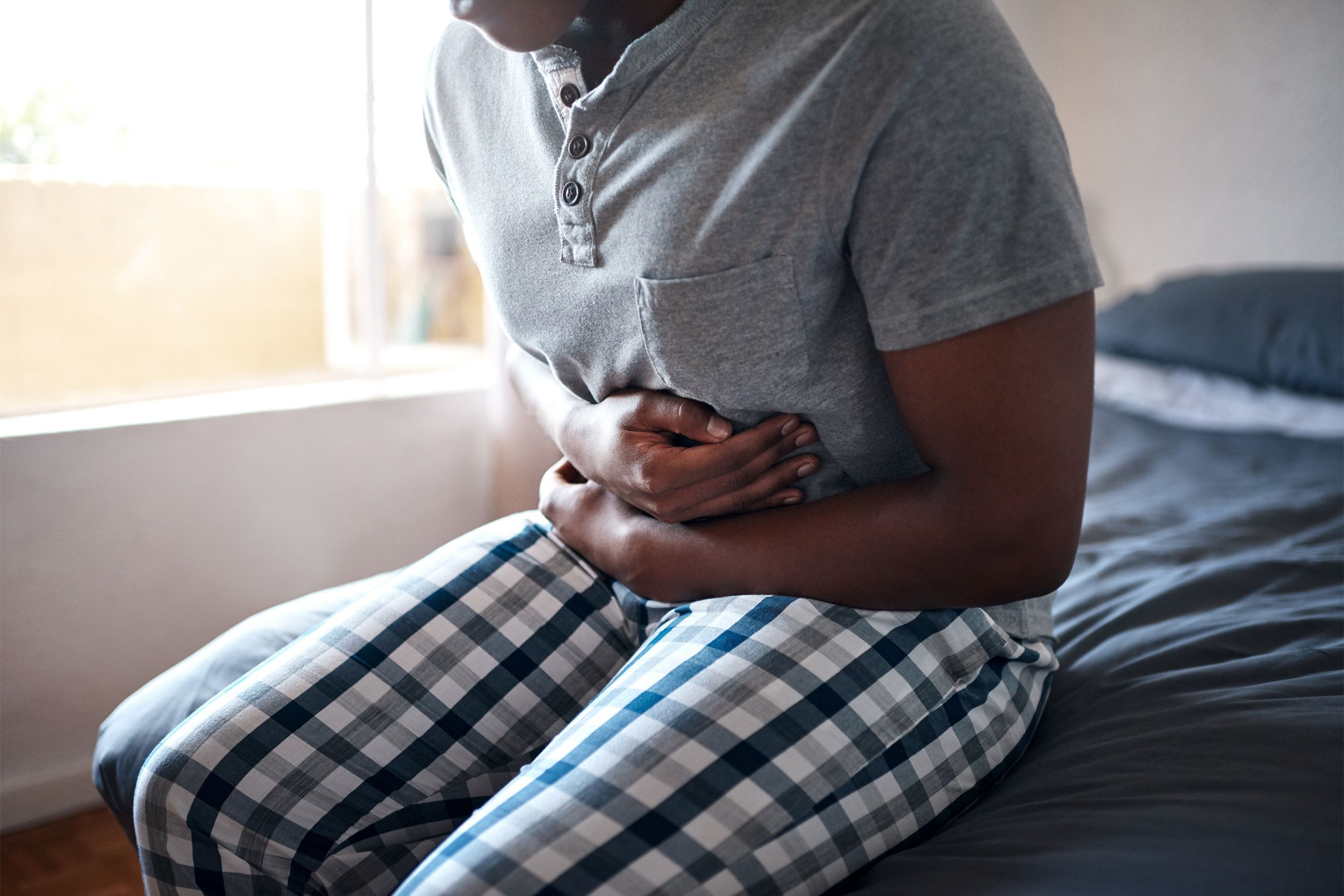 Sick man crunched over holding his stomach in pain, while sitting on the edge of the bed, wearing plaid pajama pants and a grey pajama shirt in the bedroom with windows open