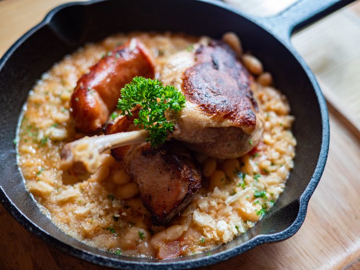 Trio Cassoulet of duck confit, pork belly and smoked sausage on a bed of white beans in a black cast iron pan.