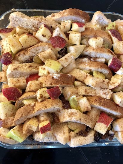 Image of glass baking dish containing prepared ingredients for a layered bread and butter pudding, slices of buttered white bread layers sprinkled with dried fruit and diced red and green apples