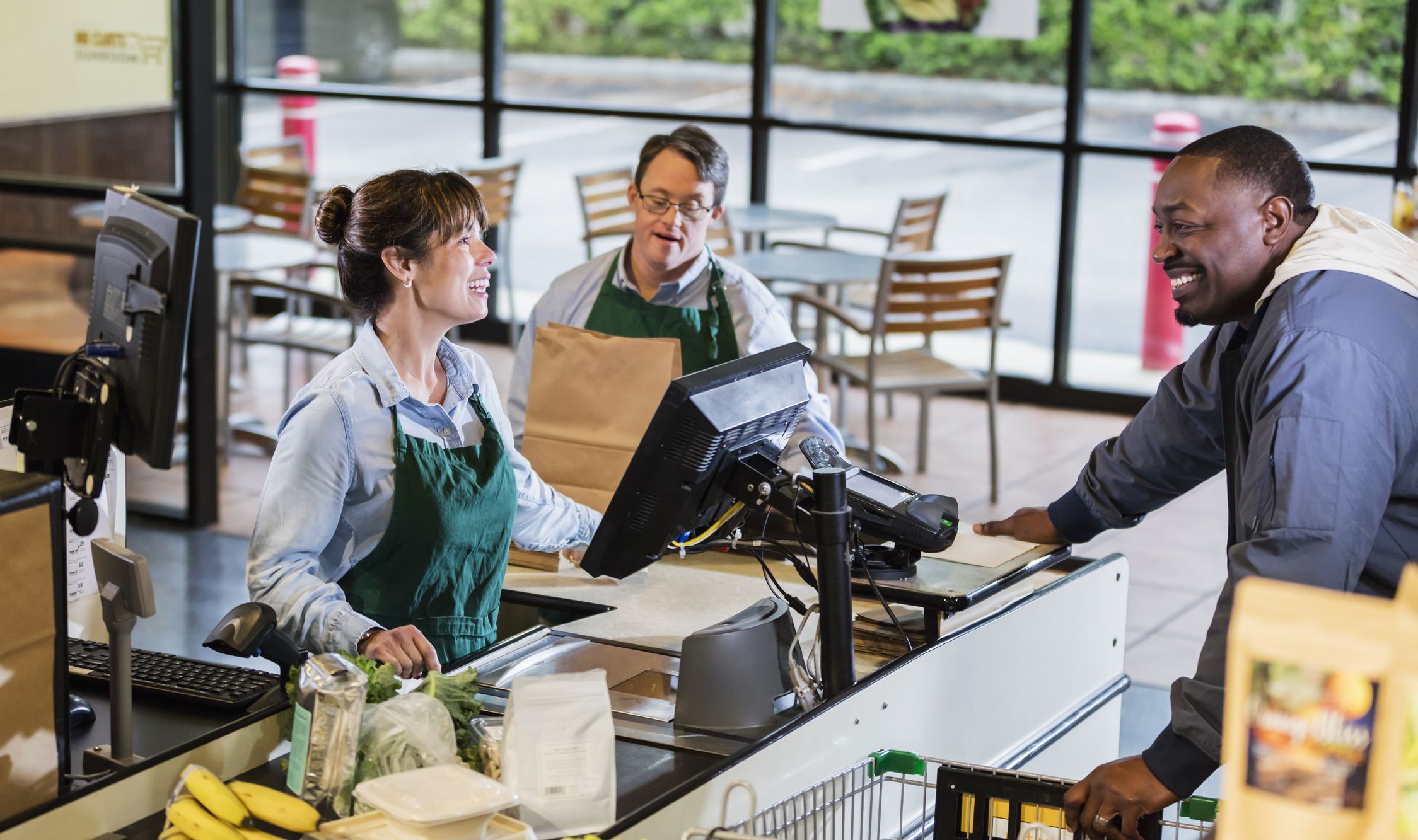 A customer and two employees at a supermarket checkout lane. A mixed race woman is the cashier and an African-American man is the shopper. They are smiling and conversing. In the background is a man with down syndrome, bagging the groceries.