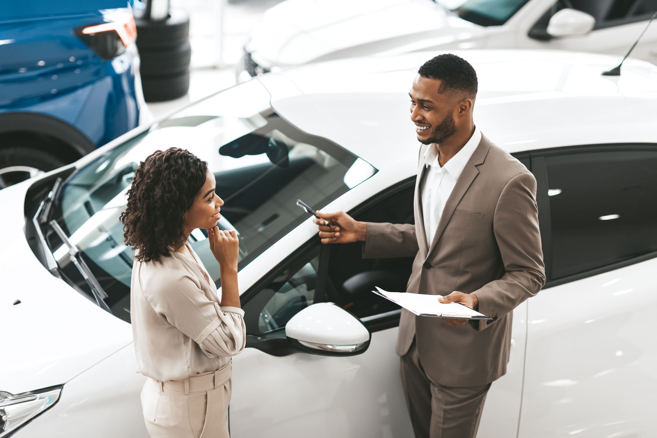 Car Sales Manager Showing Auto To Afro Lady Buyer Standing In Luxury Automobile Dealership Store. Buying Vehicle Concept