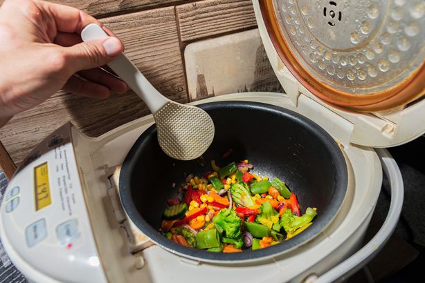 mixed set of different vegetarian vegetables in a white multicooker is stirred with a kitchen plastic Scraper. Onions, peppers, paprika, peas, corn, broccoli, cabbage and cucumber