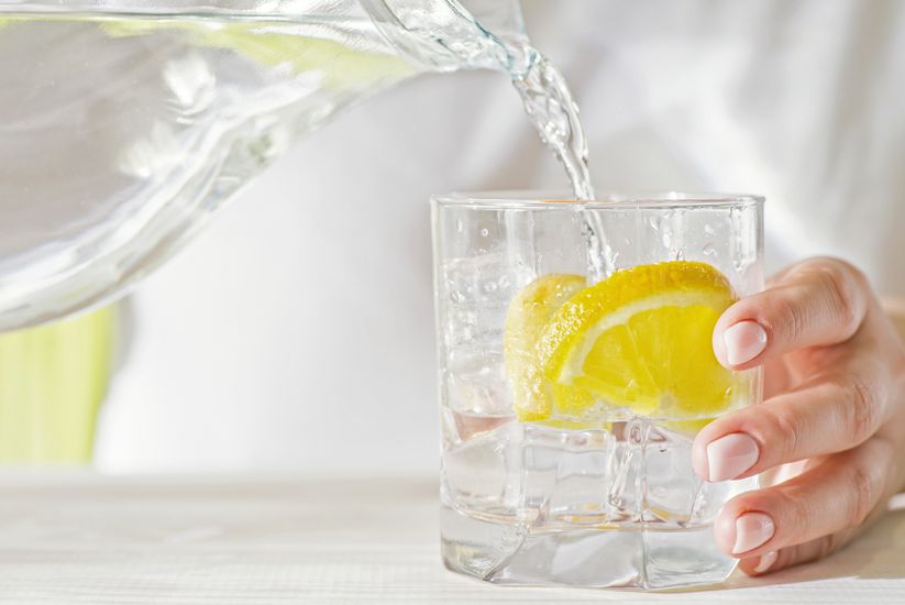 Female hands pouring water from the decanter into a glass beaker with lemon and ice. Health and diet concept. Quenching thirst on a hot day.