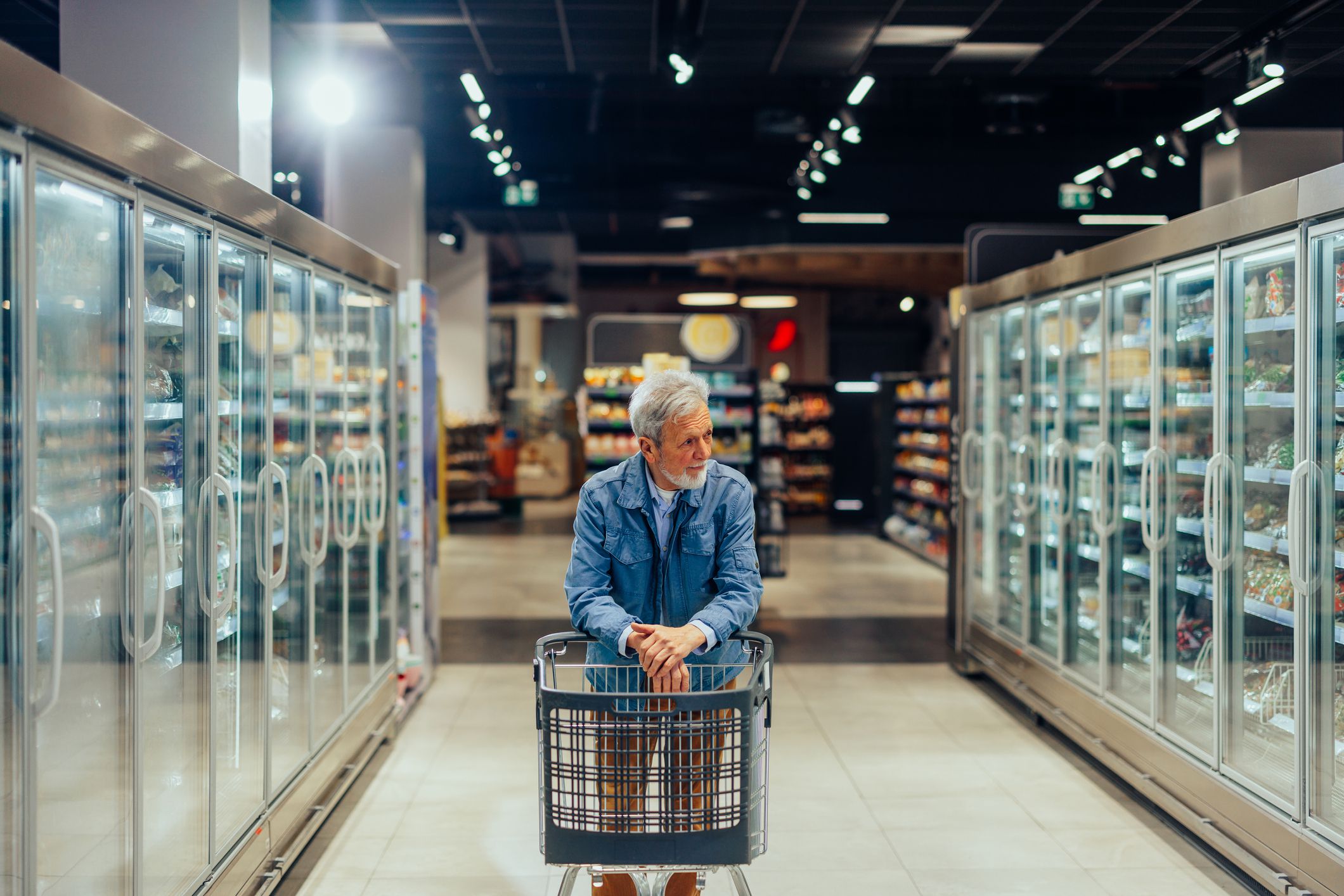 Senior man pushing a shopping cart between freezers in the grocery store and looking at the products
