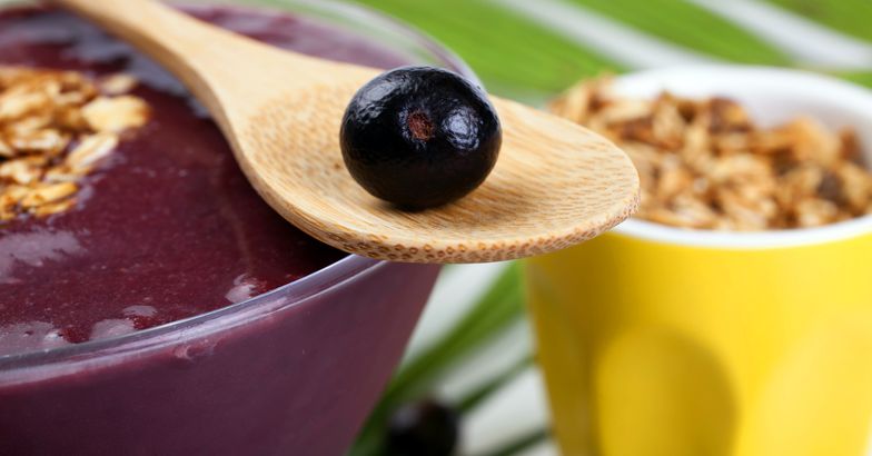 acai bowl with granola on top, glass of granola in background, wooden spoon with acai berry in foreground