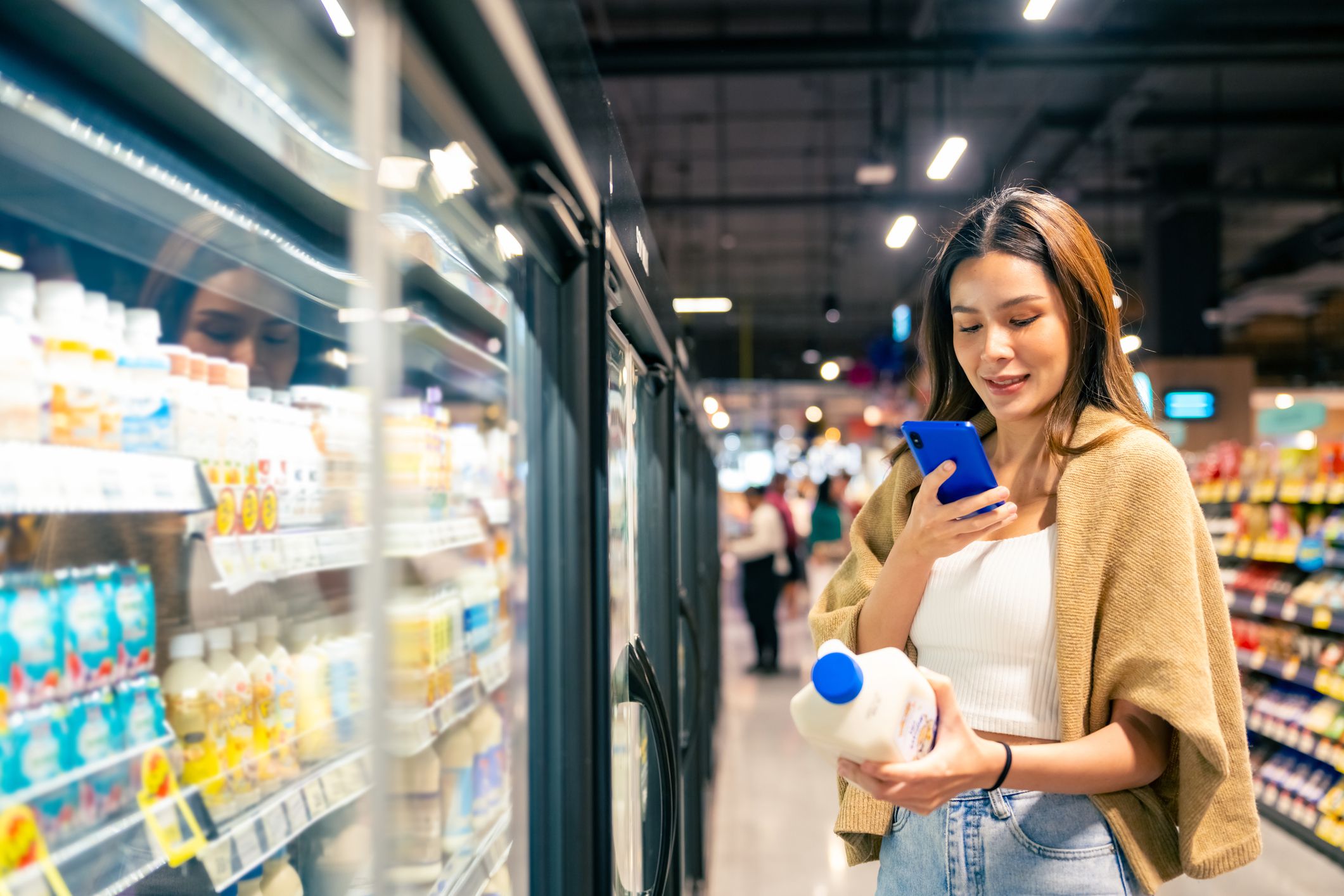 Happy Asian woman using mobile phone, shopping buying food and grocery at supermarket. Customer using smartphone scanning bar code on dairy products checking price, purchasing and making payment.