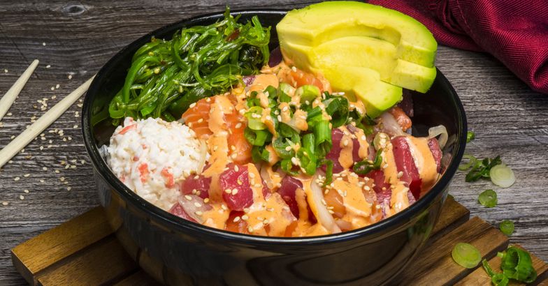 poke bowl with raw fish, avocado, sesame seeds, and algae in wooden surface with chopsticks and napkin in background